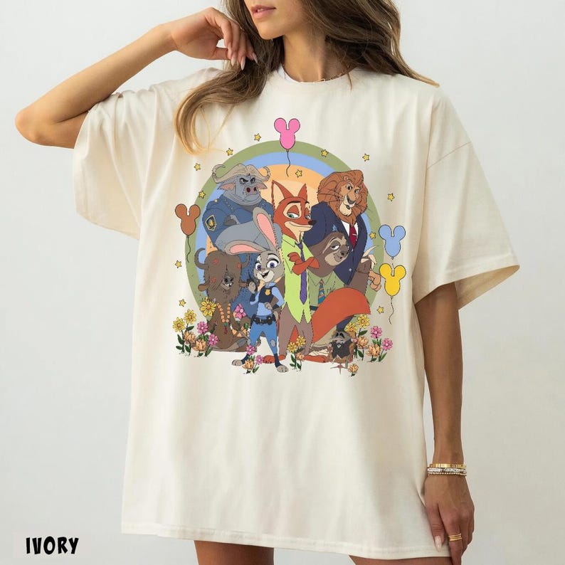 May include: Ivory-colored t-shirt featuring a colorful graphic of animated animal characters. The design includes a fox, a bunny, a lion, and other characters, set against a rainbow and floral backdrop. The word "IVORY" is printed at the bottom.