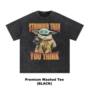 Pode incluir: T-shirt preto lavado com um gr&aacute;fico de um personagem alien&iacute;gena verde e o texto "STRONGER THAN YOU THINK" em laranja e amarelo. O personagem usa uma t&uacute;nica castanha. &Eacute; uma t-shirt lavada premium.