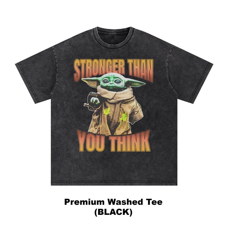 Pode incluir: T-shirt preto lavado com um gr&aacute;fico de um personagem alien&iacute;gena verde e o texto "STRONGER THAN YOU THINK" em laranja e amarelo. O personagem usa uma t&uacute;nica castanha. &Eacute; uma t-shirt lavada premium.