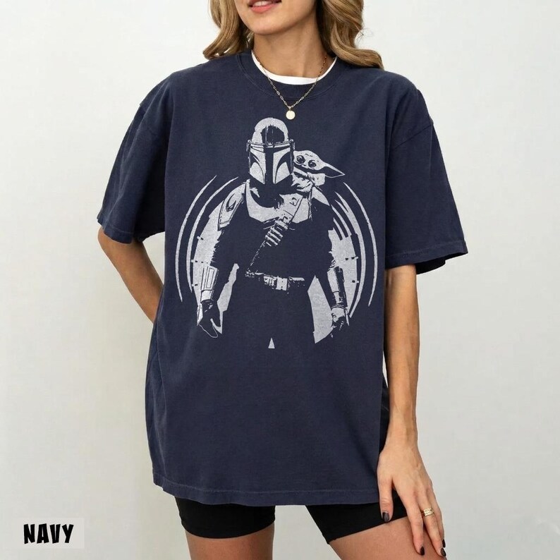 May include: Navy blue t-shirt with a white graphic of a character from a popular science fiction series and a child-like figure. The word "NAVY" is printed at the bottom. The shirt has short sleeves and a relaxed fit.