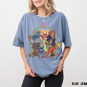 May include: A light blue t-shirt featuring a colorful graphic of characters from the animated movie Zootopia. The design includes Judy Hopps, Nick Wilde, and other characters, surrounded by flowers and Mickey Mouse-shaped balloons. The text "BLUE JEAN" is at the bottom.