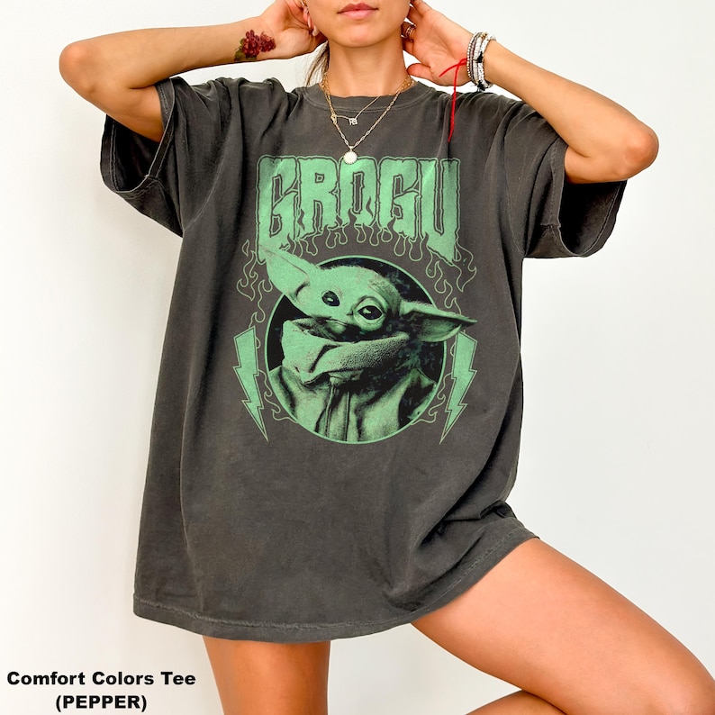 May include: Dark grey Comfort Colors tee featuring a green graphic of Grogu, with the word "GROGU" above the character. The design includes green lightning bolts and flames. The shirt is a relaxed fit.