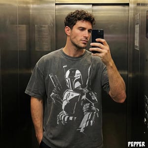 May include: A dark gray t-shirt featuring a white graphic of a character from a science fiction film. The design includes a helmet, weapons, and other details. The shirt has short sleeves and a relaxed fit.
