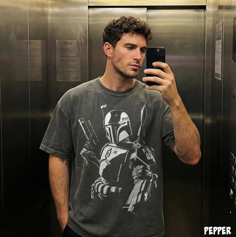 May include: A dark gray t-shirt featuring a white graphic of a character from a science fiction film. The design includes a helmet, weapons, and other details. The shirt has short sleeves and a relaxed fit.