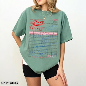 Puede incluir: Camiseta verde claro con el logo de Pizza Planet y un dise&ntilde;o de recibo. El gr&aacute;fico incluye el texto "Freshest in the Galaxy" y notas manuscritas. La camiseta es de estilo casual y de manga corta.