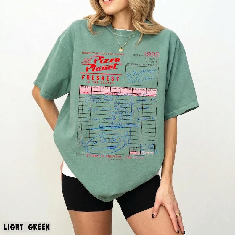 Puede incluir: Camiseta verde claro con el logo de Pizza Planet y un dise&ntilde;o de recibo. El gr&aacute;fico incluye el texto "Freshest in the Galaxy" y notas manuscritas. La camiseta es de estilo casual y de manga corta.