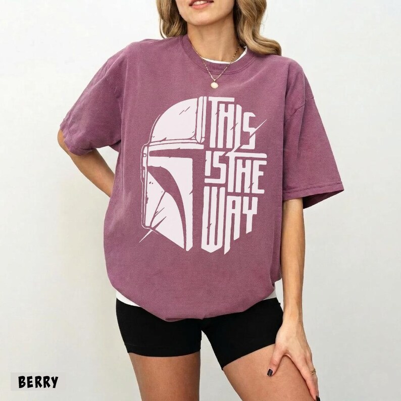 May include: Berry-coloured t-shirt with a white graphic of a helmet and the text "THIS IS THE WAY." The shirt has short sleeves and a crew neck. The model is wearing black shorts.
