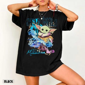 May include: Black t-shirt with a colourful graphic of Grogu from The Mandalorian, with the text "STAR WARS" and "the Mandalorian". The design includes a spaceship and other characters. The word "BLACK" is printed at the bottom.