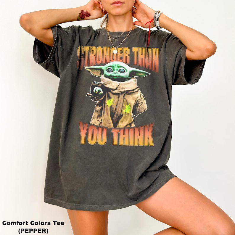 Pode incluir: T-shirt cinza escuro Comfort Colors com um gr&aacute;fico de Grogu de The Mandalorian. A imagem mostra Grogu com o texto "STRONGER THAN YOU THINK" em laranja e amarelo. A camisa tem um ajuste folgado.