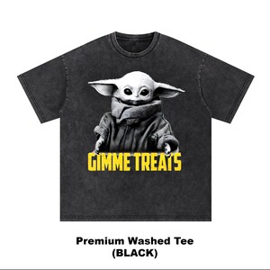 Op de afbeelding: Zwart gewassen T-shirt met een afbeelding van een personage met grote oren en de tekst "GIMME TREATS" in geel. Het personage draagt een jas en heeft een neutrale uitdrukking. Het shirt heeft een ronde hals.