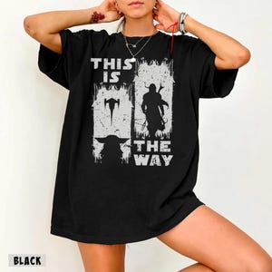 May include: Black t-shirt with a distressed white graphic featuring the text "THIS IS THE WAY" and silhouettes of a character and a creature. The shirt has a crew neck and short sleeves.