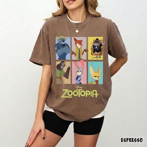 May include: Espresso-colored t-shirt featuring a colorful graphic of characters from the Disney movie Zootopia. The design includes images of a police officer, a fox, a mouse, a sloth, a rabbit, and a fennec fox. The word "Zootopia" is printed in green.