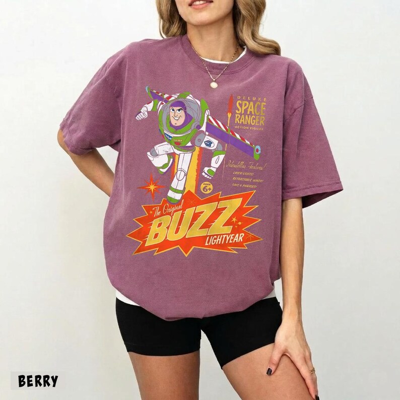 May include: A berry-colored short-sleeve t-shirt featuring a graphic of Buzz Lightyear in a dynamic pose. The print includes the text "Deluxe Space Ranger" and "The Original Buzz Lightyear" set against a large orange starburst design.