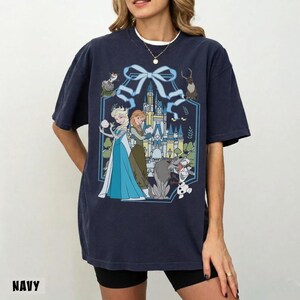 May include: Navy blue t-shirt featuring a graphic of Elsa and Anna from Frozen, with a castle backdrop and other characters. The design is framed by a blue ribbon. The word "NAVY" is printed at the bottom.
