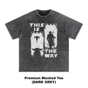 May include: Dark grey t-shirt with a white graphic featuring a silhouette of a character and the text "THIS IS THE WAY". The shirt also has an image of a spaceship and a small creature. The shirt is labeled "Premium Washed Tee (DARK GREY)".