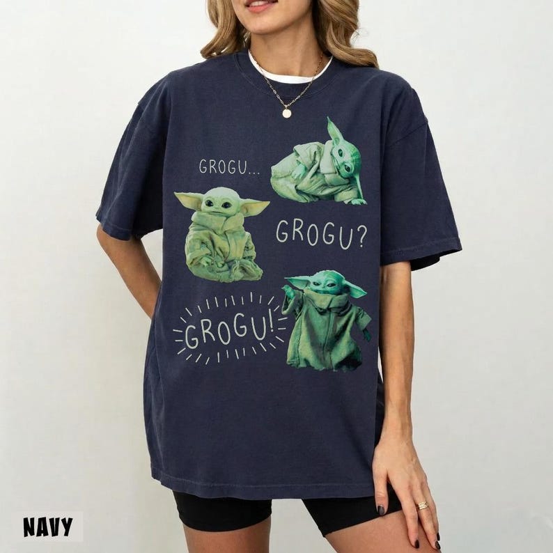 May include: Navy blue t-shirt featuring Grogu from The Mandalorian. The shirt displays Grogu in various poses with the phrases "GROGU...", "GROGU?", and "GROGU!". The word "NAVY" is printed at the bottom left.