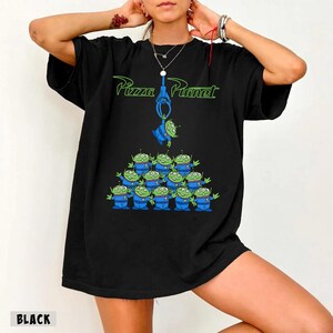 May include: Black t-shirt featuring the Pizza Planet logo in green and a graphic of the Little Green Men from Toy Story. The design includes a claw machine and a pyramid of the aliens. The word "BLACK" is printed at the bottom.