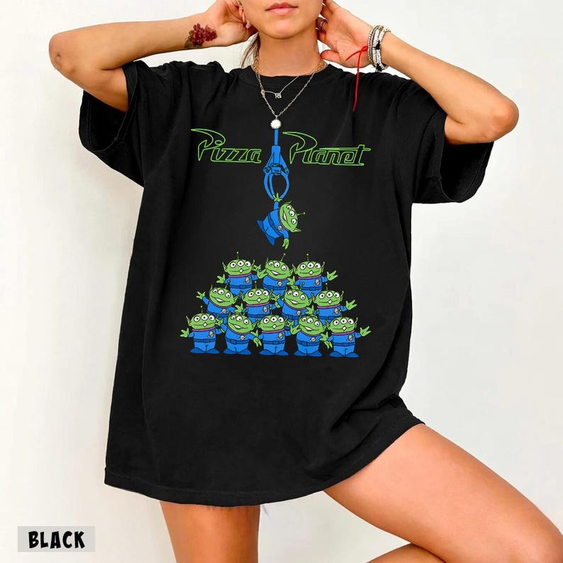 May include: Black t-shirt featuring the Pizza Planet logo in green and a graphic of the Little Green Men from Toy Story. The design includes a claw machine and a pyramid of the aliens. The word "BLACK" is printed at the bottom.