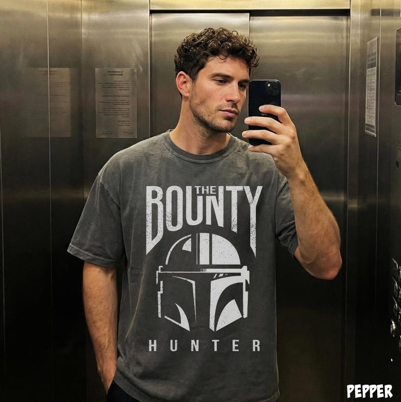 Puede incluir: Camiseta gris oscuro con el texto "THE BOUNTY HUNTER" y un gr&aacute;fico blanco de un casco. La camiseta tiene un aspecto descolorido. El dise&ntilde;o est&aacute; centrado en la parte delantera de la camiseta.