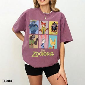 May include: Berry-colored t-shirt featuring a colorful graphic of characters from the Disney movie Zootopia. The design includes images of a police officer, a fox, a weasel, a sloth, a rabbit, and a fennec fox. The word "Zootopia" is printed in green.