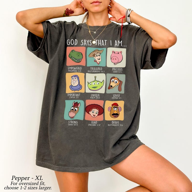 May include: Dark gray t-shirt with the text "GOD SAYS THAT I AM..." and cartoon character illustrations. The shirt features the words "Empowered," "Treasured," "Precious," "Important," "Unique," "Loved," "Strong," "Kind," and "Brave."