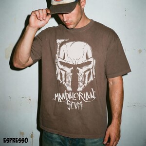 Pu&ograve; includere: T-shirt color espresso con grafica bianca di un casco a forma di teschio e la scritta "MANDALORIAN SCUM". La maglietta girocollo a maniche corte &egrave; di colore marrone scuro.