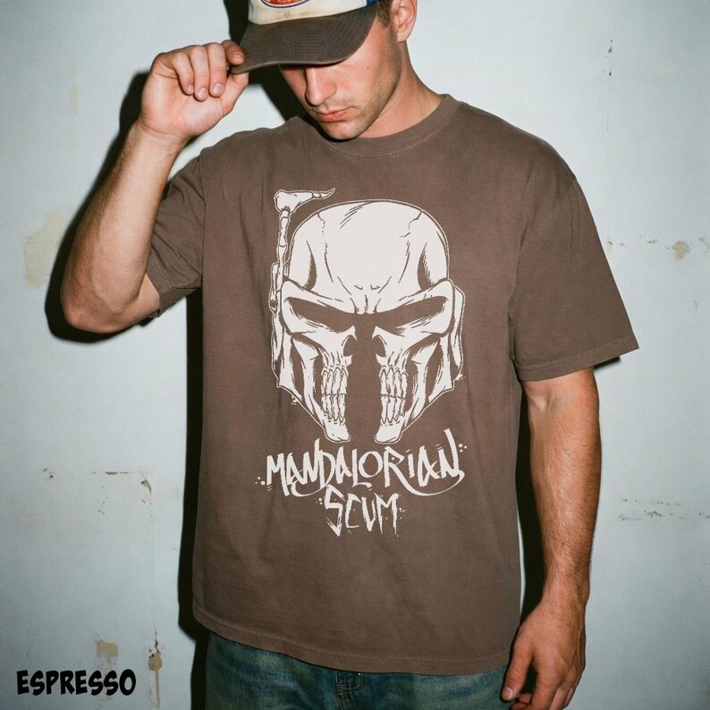 Pu&ograve; includere: T-shirt color espresso con grafica bianca di un casco a forma di teschio e la scritta "MANDALORIAN SCUM". La maglietta girocollo a maniche corte &egrave; di colore marrone scuro.