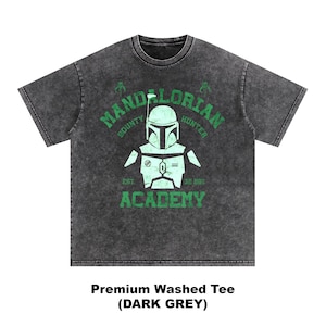 Peut inclure: T-shirt gris fonc&eacute; d&eacute;lav&eacute; avec un motif vert. Le motif comprend les mots "MANDALORIAN BOUNTY HUNTER ACADEMY" et une illustration d'un casque et d'une armure Mandaloriens. Le t-shirt est &eacute;tiquet&eacute; "Premium Washed Tee (DARK GREY)".
