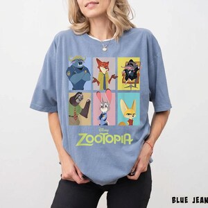 May include: A light blue t-shirt featuring a colorful graphic of characters from the Disney movie Zootopia. The design includes images of a buffalo, fox, mouse, sloth, rabbit, and fennec fox. The word "Zootopia" is printed in green.