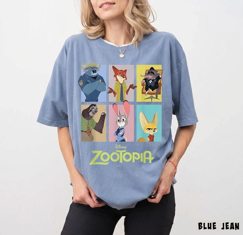 May include: A light blue t-shirt featuring a colorful graphic of characters from the Disney movie Zootopia. The design includes images of a buffalo, fox, mouse, sloth, rabbit, and fennec fox. The word "Zootopia" is printed in green.