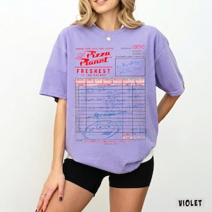 Puede incluir: Camiseta violeta con un dise&ntilde;o de recibo de Pizza Planet. El gr&aacute;fico incluye el logotipo de Pizza Planet, el texto "FRESHEST IN THE GALAXY" y detalles manuscritos. La camiseta tiene mangas cortas y un corte holgado.