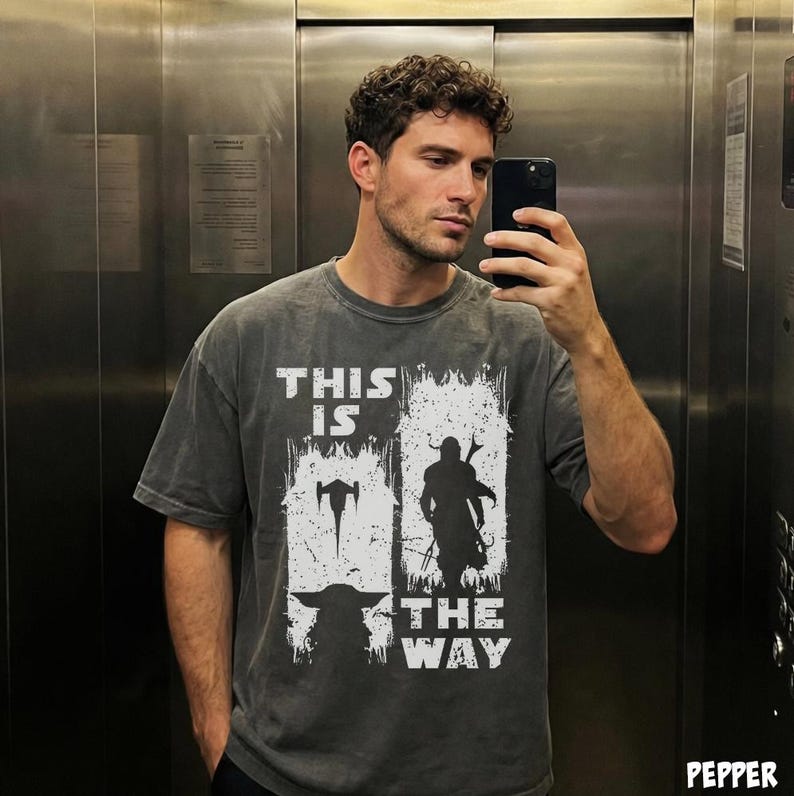 May include: Dark gray t-shirt with the text "THIS IS THE WAY" in white. The graphic features a silhouette of a character and a baby-like creature. The shirt is a short-sleeved, crew neck style.
