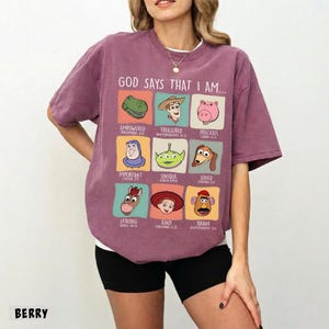 May include: Berry-colored t-shirt with the text "GOD SAYS THAT I AM..." and cartoon character graphics. The shirt features a crew neck and short sleeves. The graphic includes the words "Empowered", "Treasured", "Precious", "Important", "Unique", "Loved", "Strong", "Kind", and "Brave".
