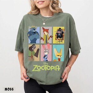 May include: A moss green t-shirt featuring a grid of six colorful character portraits from the Disney movie Zootopia. The movie title is printed in a light green font below the portraits. The word "MOSS" is printed in the bottom left corner.