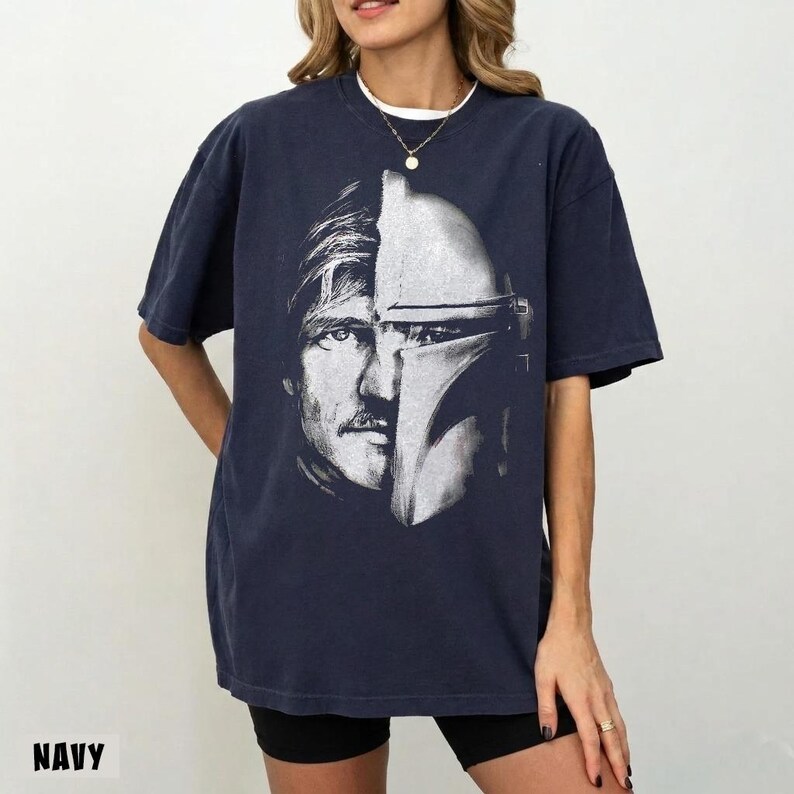 May include: Navy blue t-shirt featuring a split image of a man's face and a Mandalorian helmet. The graphic is in black and white. The word "NAVY" is printed at the bottom of the shirt.