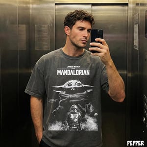 May include: Dark gray t-shirt featuring a graphic from the Star Wars series, "The Mandalorian." The design includes the text "STAR WARS THE MANDALORIAN" above an image of Grogu and the Mandalorian. The shirt has a vintage, worn look.
