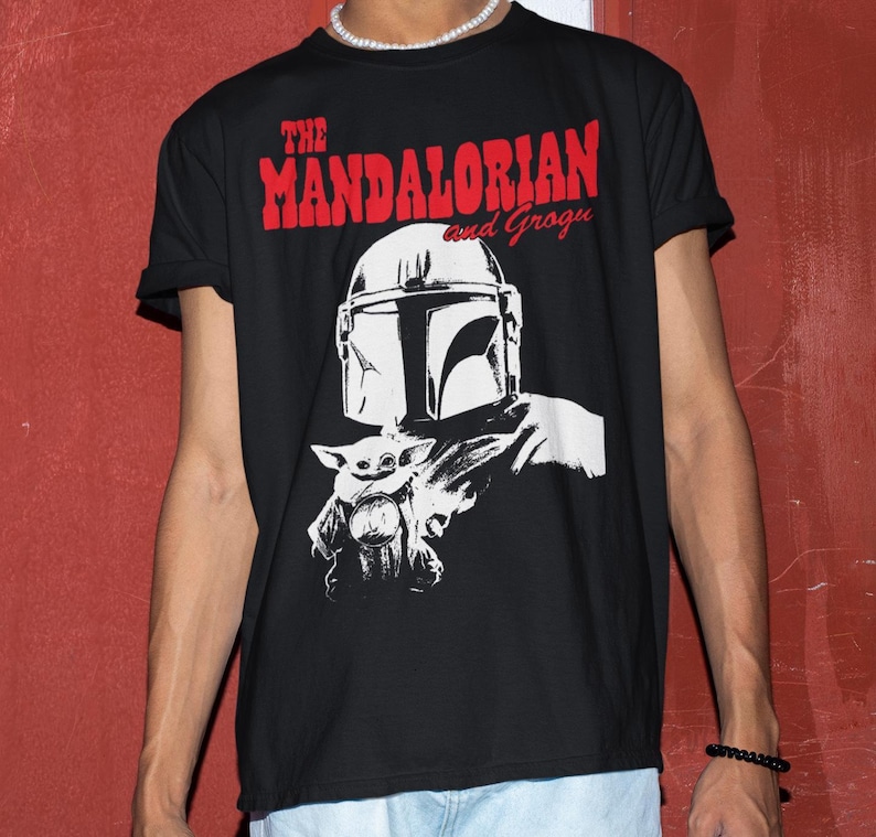 Op de afbeelding: Zwart T-shirt met een witte afbeelding van The Mandalorian en Grogu. De tekst "THE MANDALORIAN and Grogu" is in het rood boven de afbeelding gedrukt. Het ontwerp bevat de helm van de Mandalorian en Grogu.