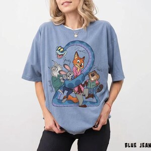 May include: A light blue t-shirt featuring a cartoon graphic of several animal characters, including a fox, rabbit, and beaver, with the number "2" formed by a blue snake. The shirt has the words "BLUE JEAN" printed at the bottom.