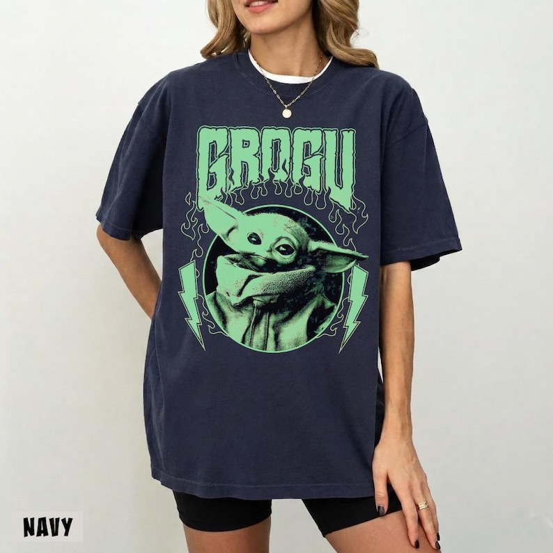 May include: Navy blue t-shirt featuring a green graphic of Grogu, with the text "GROGU" above the image. The design includes lightning bolts and a flame-like border. The shirt is a relaxed fit.