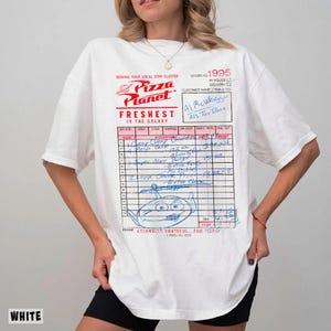 Puede incluir: Camiseta blanca con un dise&ntilde;o de recibo de Pizza Planet. El gr&aacute;fico incluye el logotipo de Pizza Planet, texto y un boceto de personaje de dibujos animados. La camiseta tiene mangas cortas y un corte holgado.