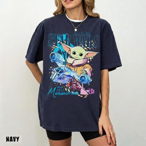 May include: Navy blue t-shirt with a graphic of Grogu from The Mandalorian, featuring the text "STAR WARS" and "The Mandalorian". The design includes a spaceship and colourful accents. The shirt has a relaxed fit.