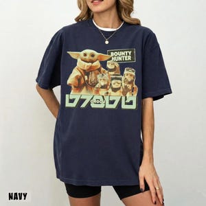 K&ouml;nnte beinhalten: Marineblaues T-Shirt mit einem Aufdruck von Grogu und anderen Star Wars-Charakteren. Das Design enth&auml;lt den Text "BOUNTY HUNTER" und "GROGU" in Gr&uuml;n. Das Shirt hat eine lockere Passform.