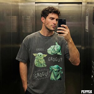 May include: A gray t-shirt featuring a graphic of Grogu from The Mandalorian. The design includes multiple images of Grogu with the text "GROGU...", "GROGU?", and "GROGU!". The shirt is a casual style.