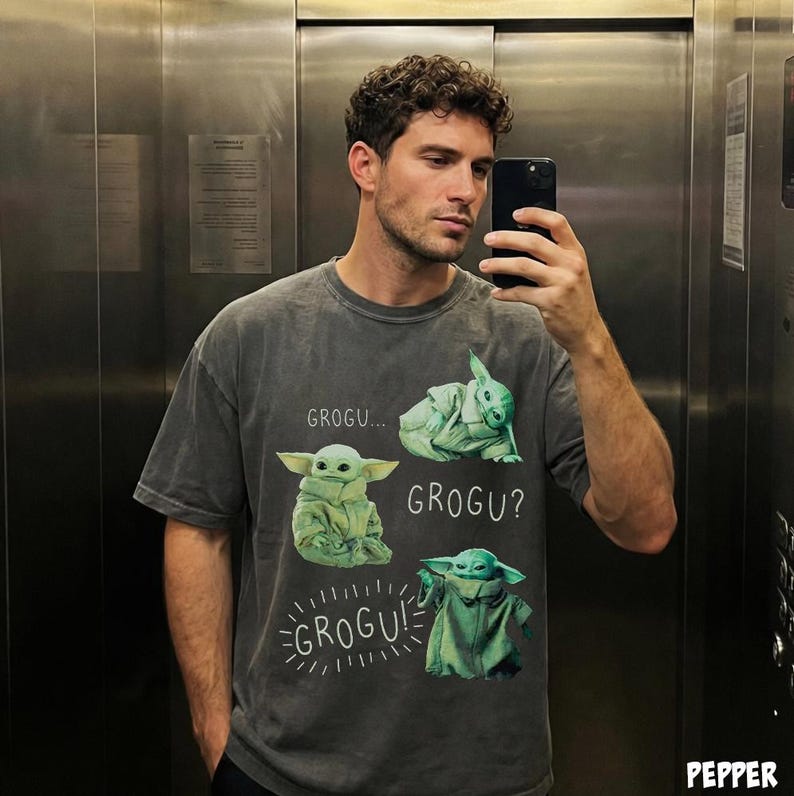 May include: A gray t-shirt featuring a graphic of Grogu from The Mandalorian. The design includes multiple images of Grogu with the text "GROGU...", "GROGU?", and "GROGU!". The shirt is a casual style.