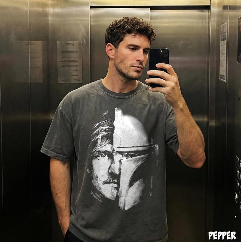 May include: A gray t-shirt featuring a split image of a character's face and helmet. The graphic is in black and white. The shirt has short sleeves and a relaxed fit.