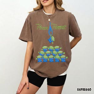 May include: Espresso-colored t-shirt featuring a graphic of green aliens from Pizza Planet. The aliens are arranged in a pyramid formation, with one being lifted by a blue claw. The words "Pizza Planet" are printed above.