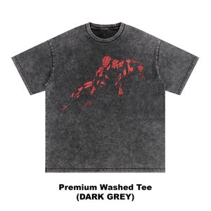 May include: A dark grey short-sleeve t-shirt with a washed, mottled texture. The center of the chest features a red graphic print of a crouching superhero figure with a webbed pattern. The text below the shirt reads Premium Washed Tee (DARK GREY).