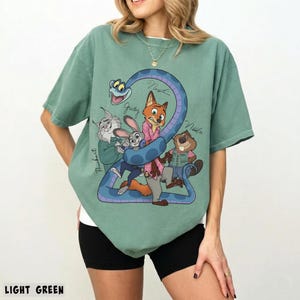 May include: Light green t-shirt featuring a cartoon graphic of several animal characters, including a fox, rabbit, and beaver, with the number 2 and the names of the characters. The text "LIGHT GREEN" is at the bottom.