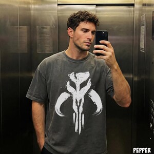 May include: A dark gray t-shirt featuring a white graphic of a skull-like design with curved horns. The shirt has a vintage, washed appearance. The person in the image is holding a black smartphone.