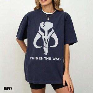 May include: Navy blue t-shirt featuring a white graphic of a skull with horns and the text "THIS IS THE WAY." The shirt is a relaxed fit and has short sleeves. The word "NAVY" is printed at the bottom.